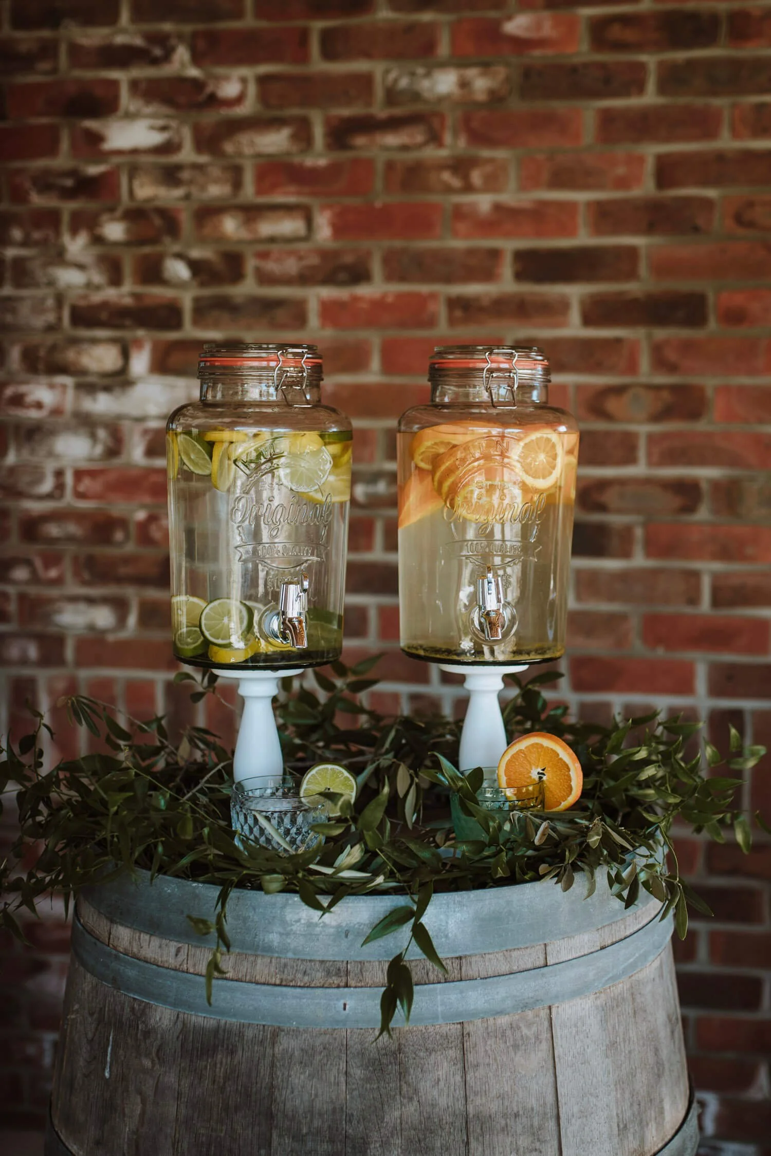 Kilner Drinks Dispensers filled with citrus water on white stands atop a wooden barrel, available to hire for your wedding or event.
