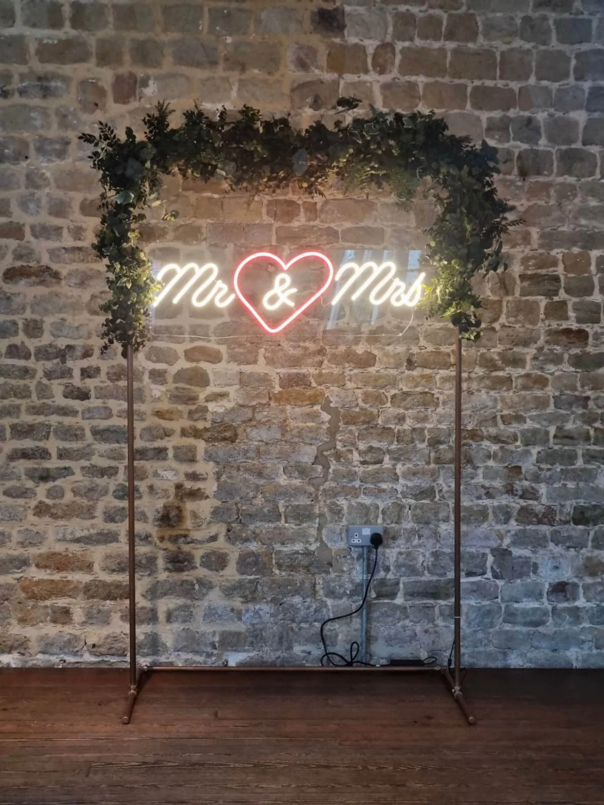 Mr & Mrs Neon Sign with white and red neon lights on a foliage-decorated stand, available to hire for your wedding or event.