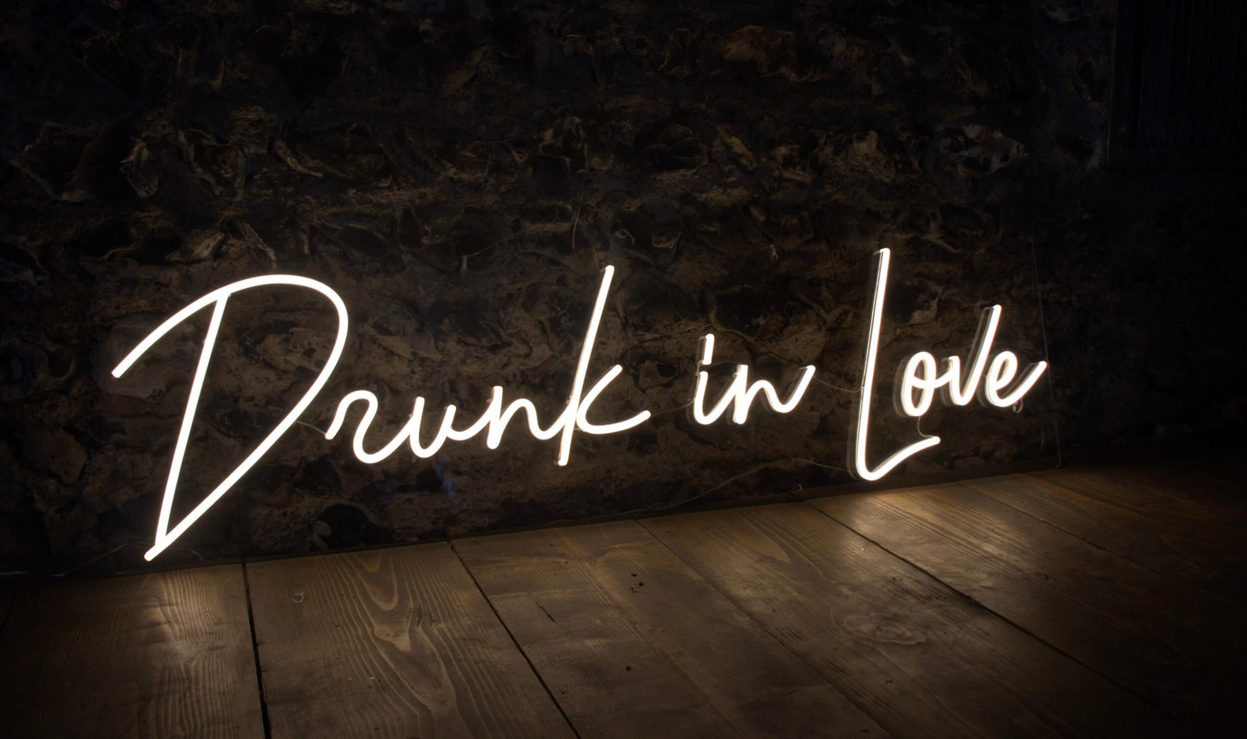 Drunk in Love Neon Sign in white neon light on a dark stone wall and wooden floor, available to hire for your wedding or event.