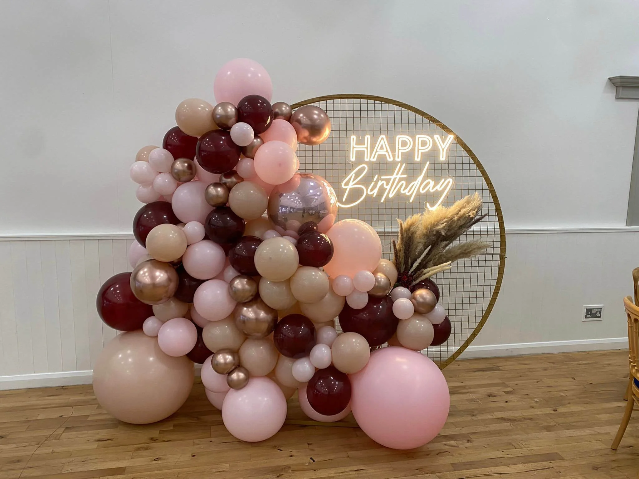 Happy Birthday Neon Sign with pink, beige, burgundy, and metallic rose gold balloons and pampas grass, available to hire for your wedding or event.