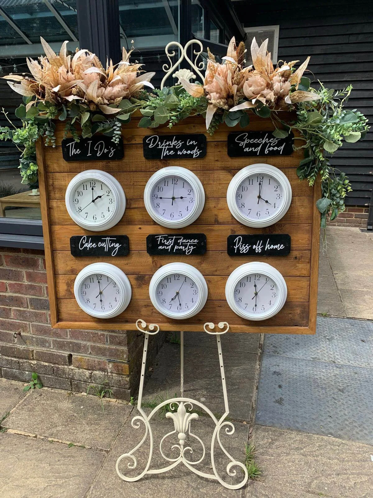 Clocks Run of the Day - 6 clocks with chalkboards on a wooden board decorated with faux greenery and beige florals, available to hire for your wedding or event.