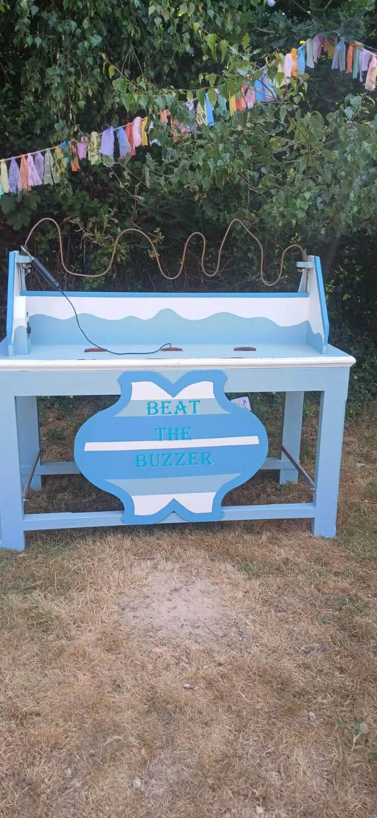 Beat the Buzzer game table in light blue and white, available to hire for your wedding or event, set outdoors with colorful bunting above.