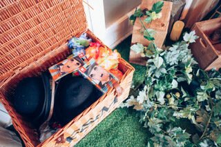Prop box containing a black hat and colorful leis, available to hire for your wedding or event, styled on green grass with faux greenery nearby.