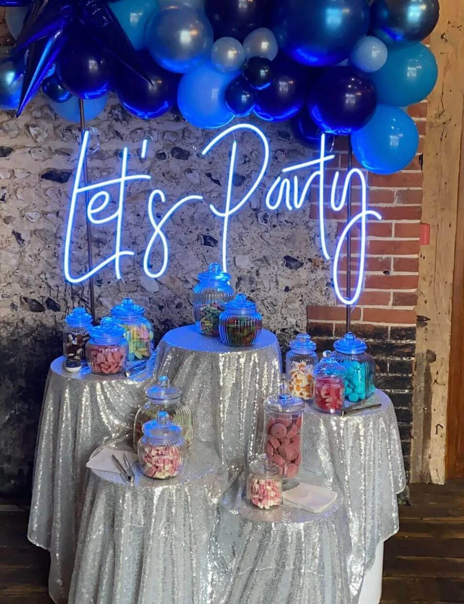 Let's Party Colour Changing Neon Sign glowing bright blue above silver sequin tables with jars of sweets and blue balloons, available to hire for your wedding or event.