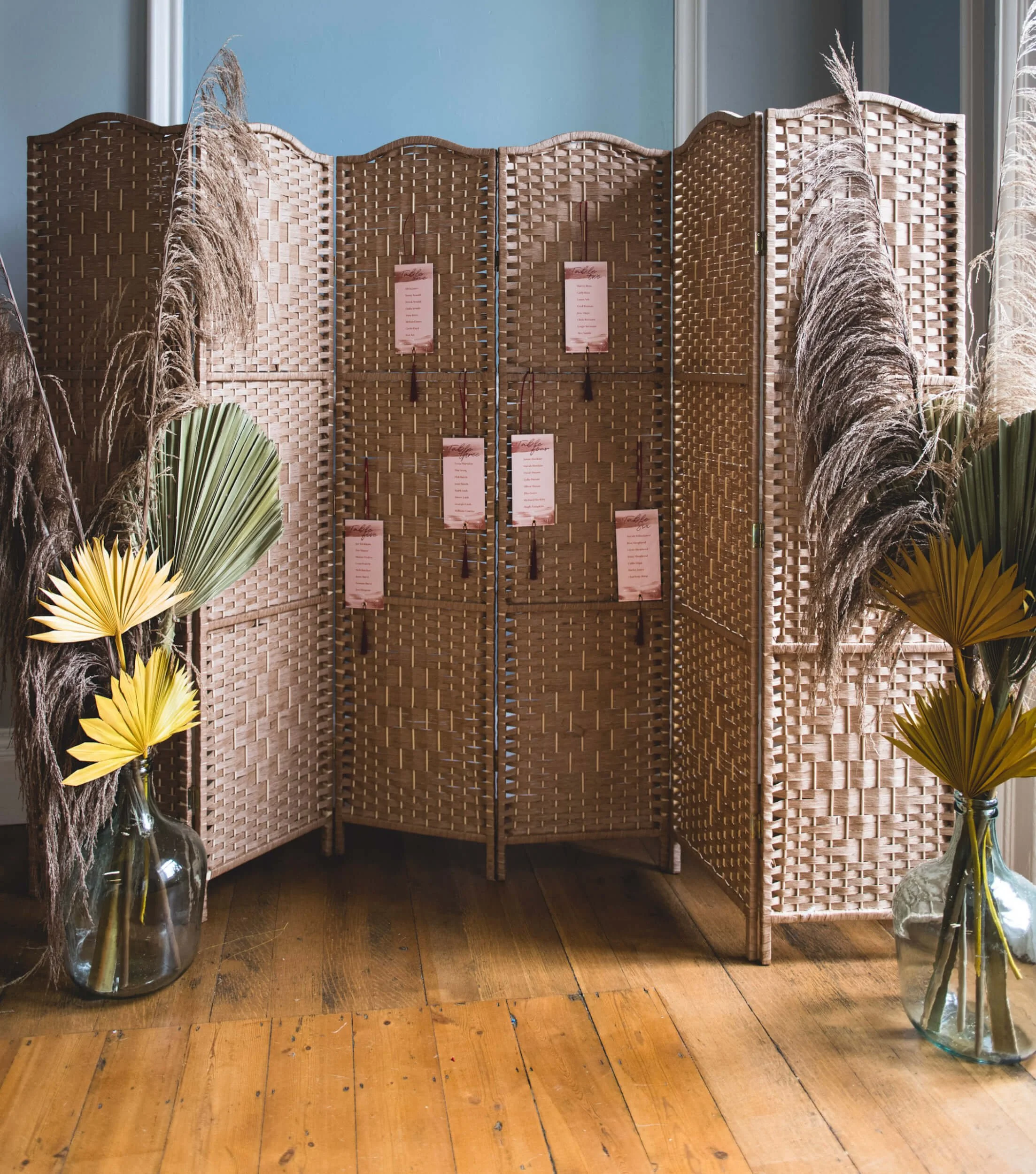 Wicker Room Divider with four natural woven panels, decorated with hanging pink cards, surrounded by dried pampas grass and yellow palm leaves, available to hire for your wedding or event.