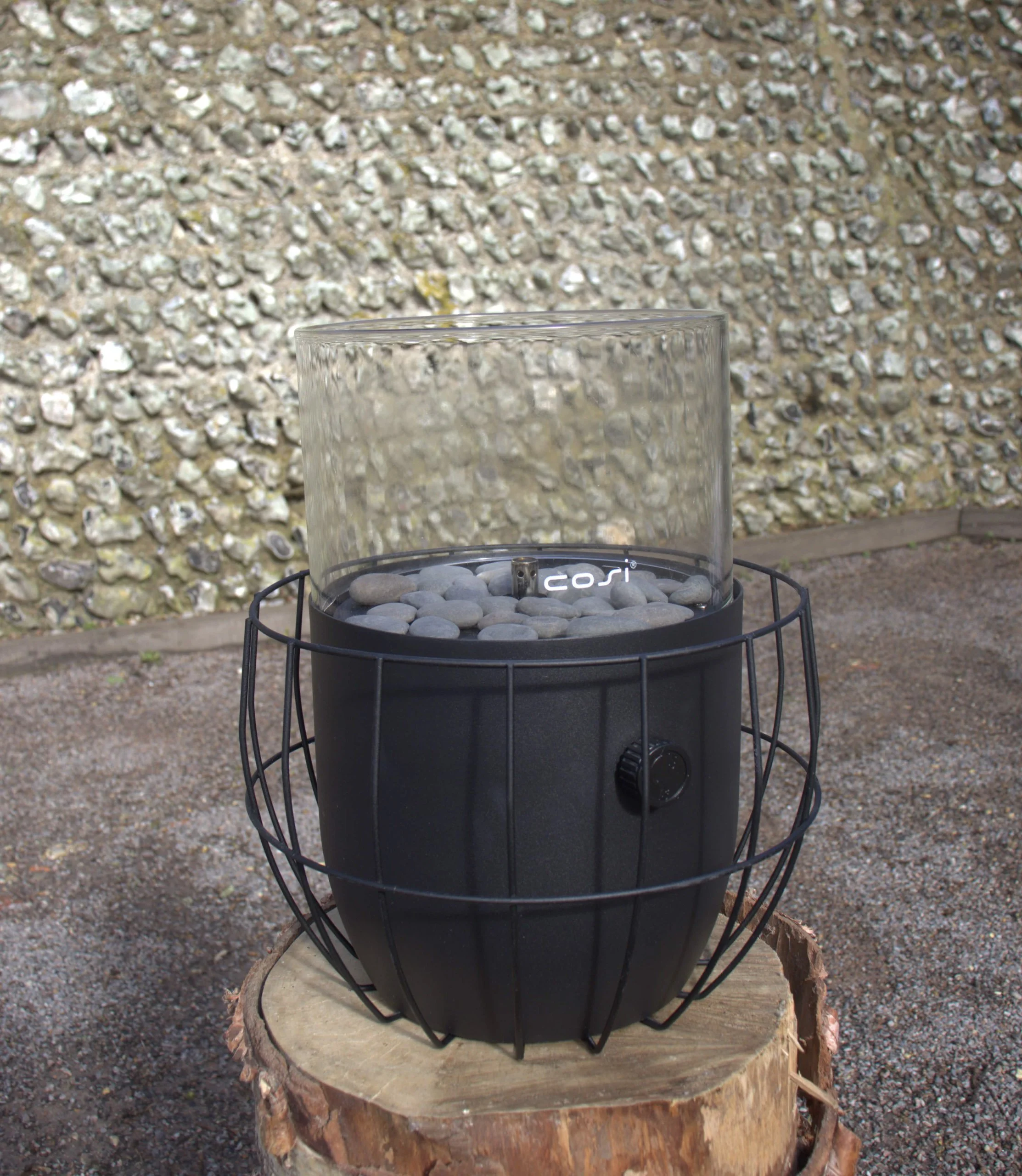 Firepit - cosipit with glass wind guard and stones, available to hire for your wedding or event, shown outdoors on a wooden log base.
