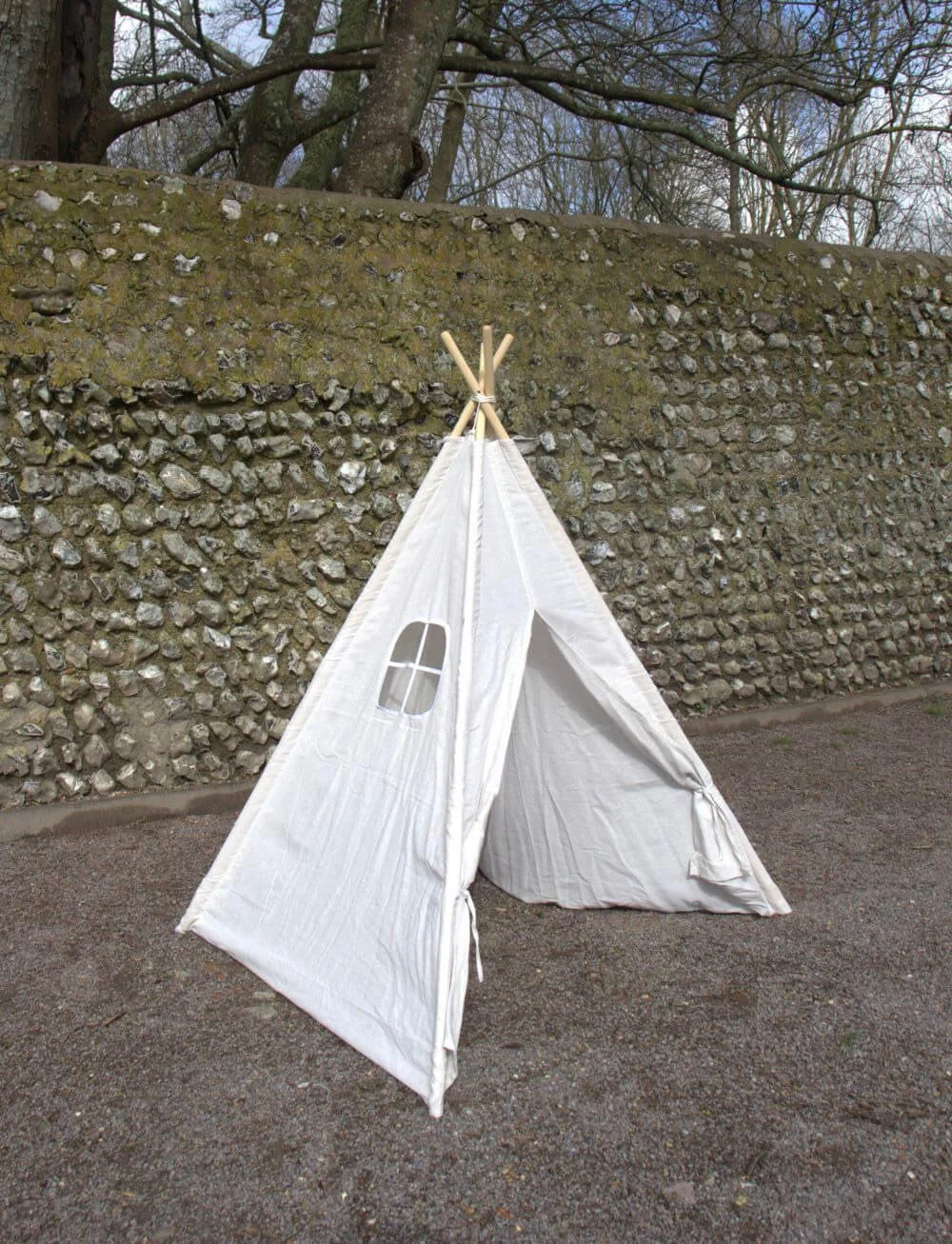 Children's Teepee Play Tent in white with a small window, available to hire for your wedding or event, set outdoors on gravel by a stone wall.