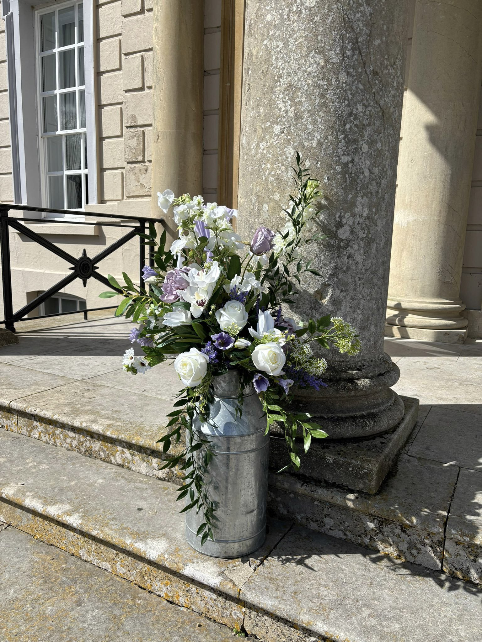 Milk Churns filled with white and purple flowers and greenery, available to hire for your wedding or event, displayed on stone steps by a column.