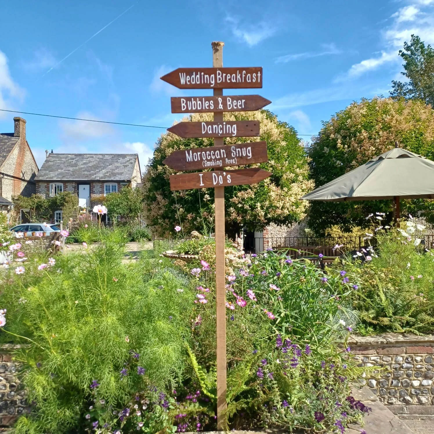 Upwaltham Festival Sign - Rustic wooden directional signpost available to hire for your wedding or event, placed in a garden setting with flowers and greenery.