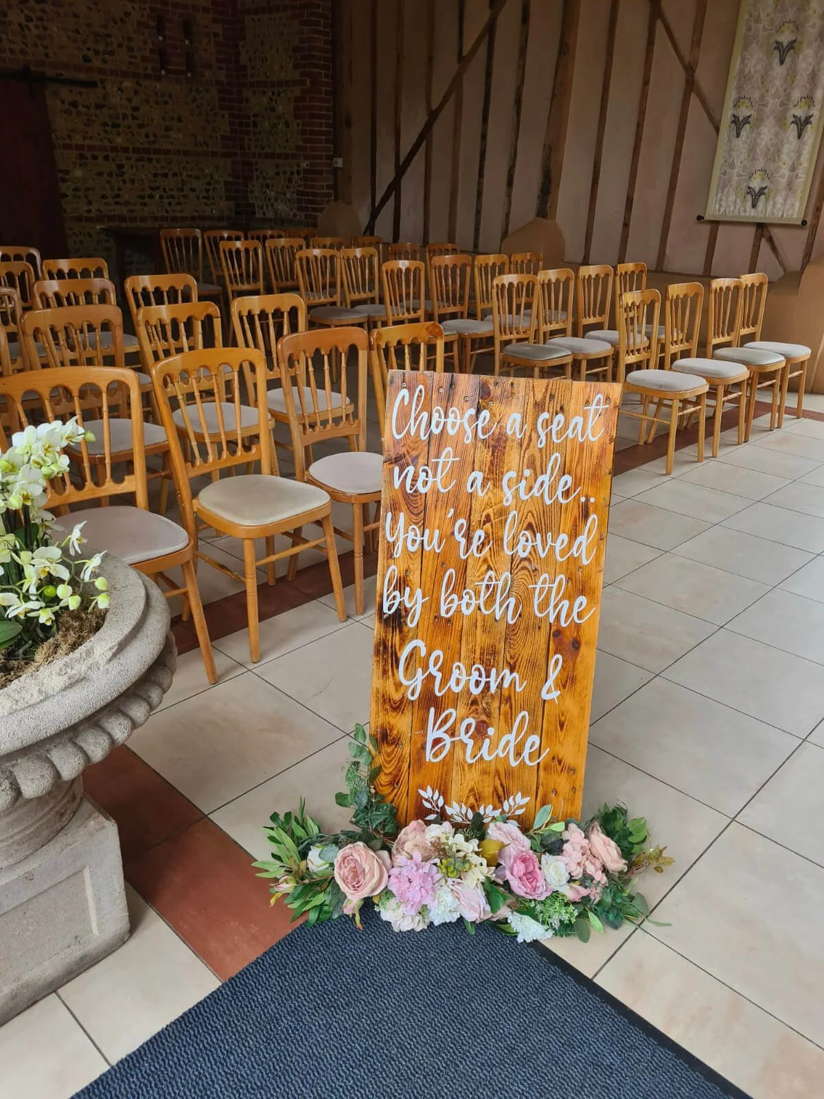 Wooden Rustic Pick A Seat Not A Side Sign - A1 with white script text and floral base, available to hire for your wedding or event.