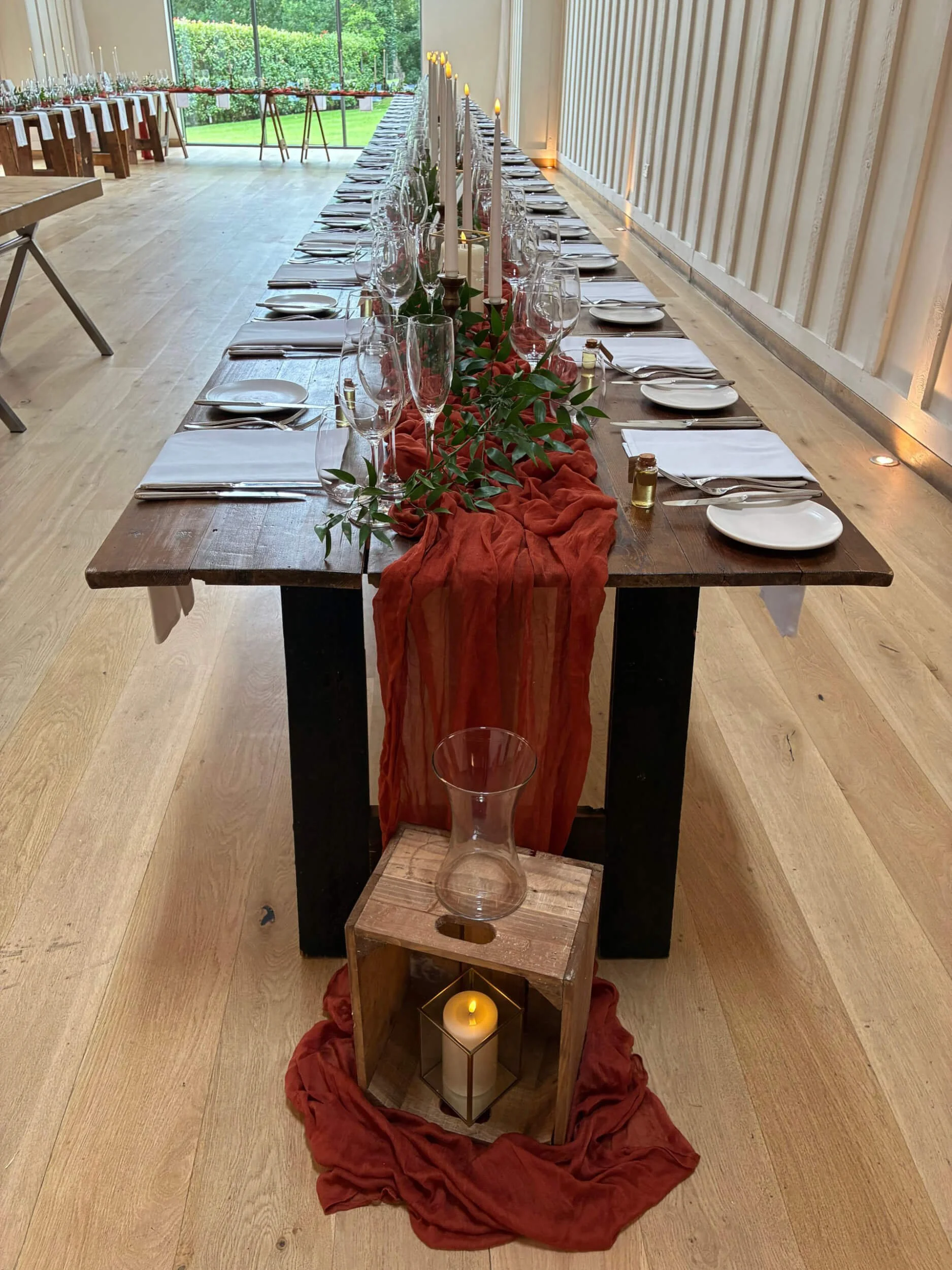 Cheesecloth Table Runner - Burnt Orange draped along a long wooden table with greenery and candles, available to hire for your wedding or event.