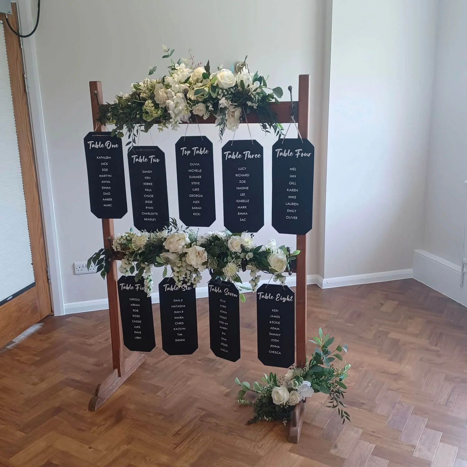Rustic Tableplan with black cards and white floral decorations on a wooden frame, available to hire for your wedding or event.