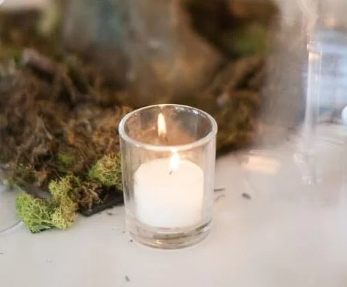 Clear Glass Tealight with a lit white candle, available to hire for your wedding or event, displayed on a white surface with moss decoration.