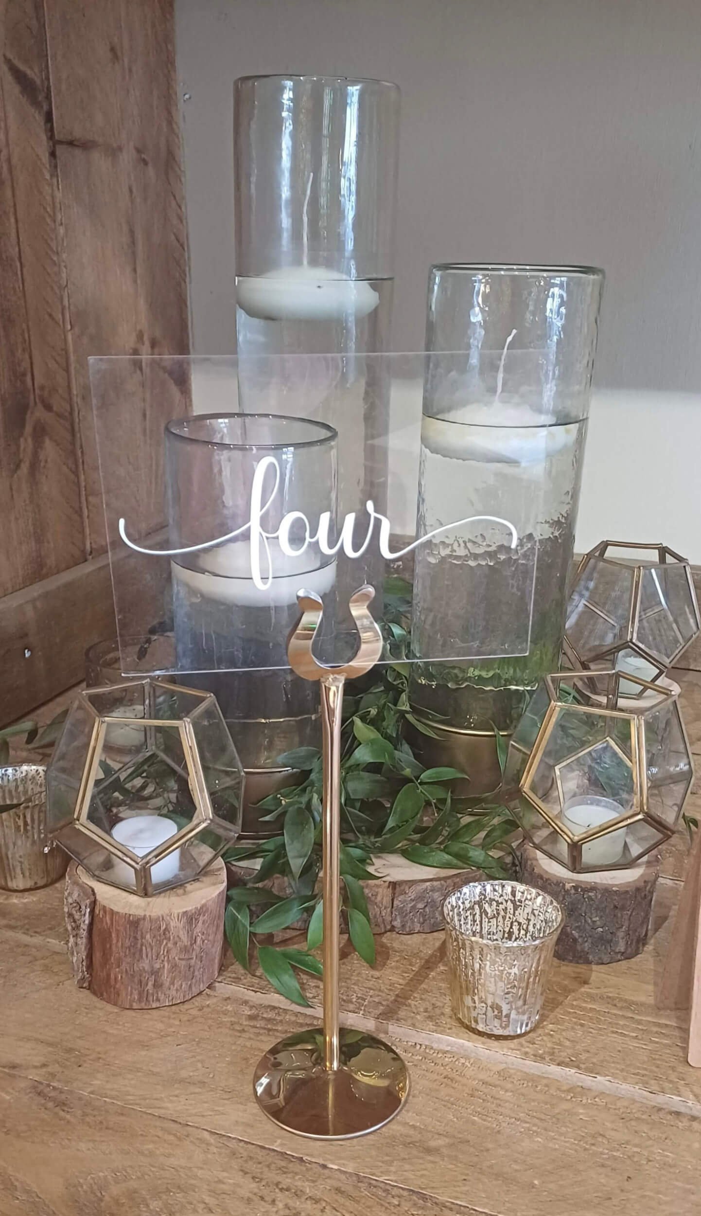 Table Number Holder - Gold with a clear acrylic card reading 'four', surrounded by glass candle holders and greenery, available to hire for your wedding or event.
