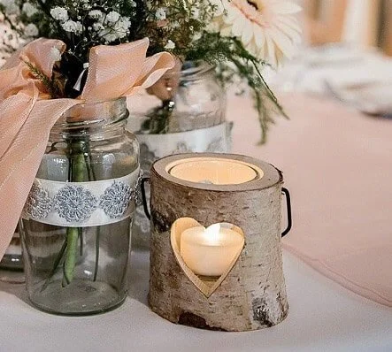 Rustic Wooden Tealight with heart cutout and lit candle, available to hire for your wedding or event, styled on a table with floral jars and pink ribbon.
