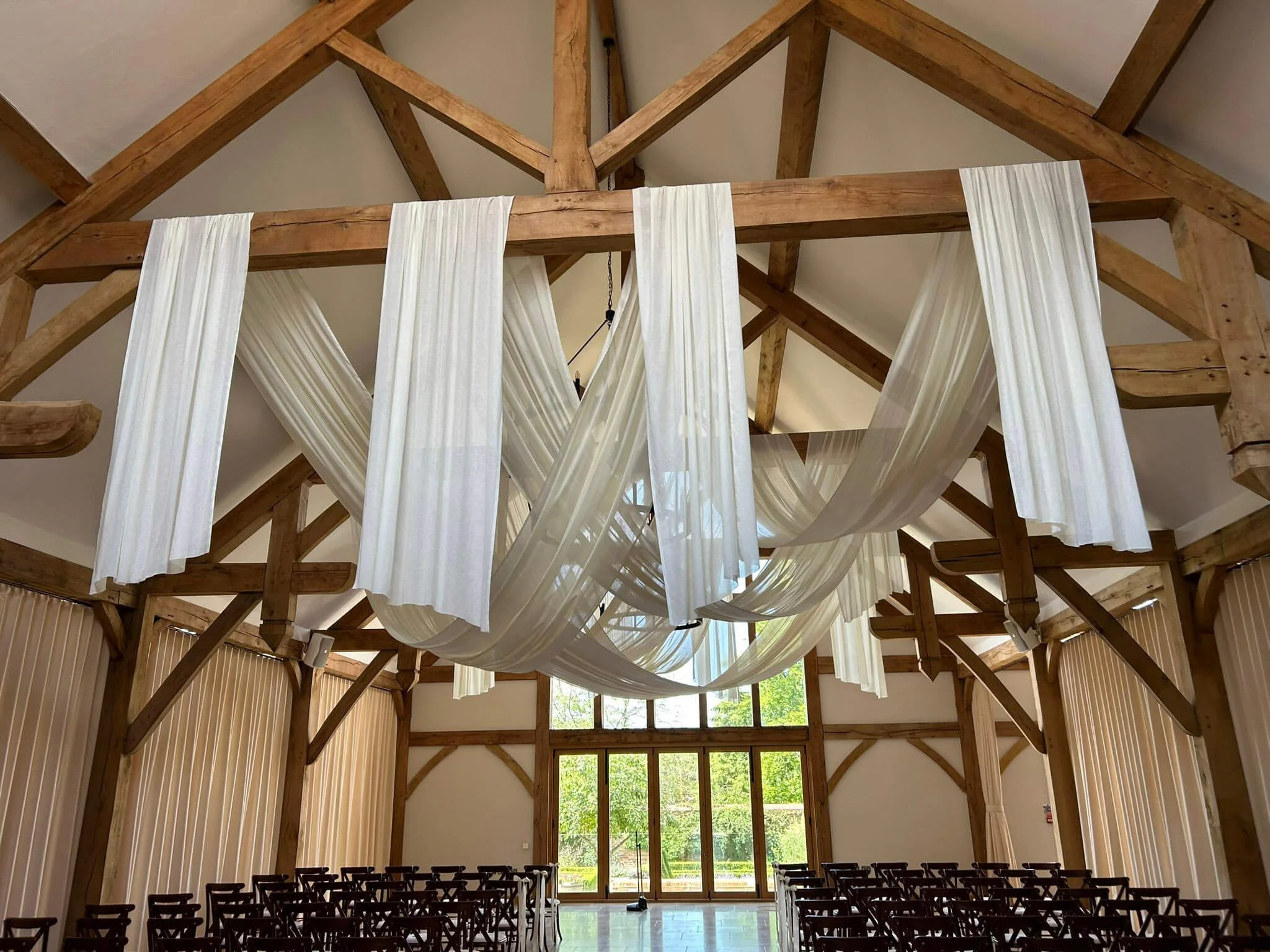 Organic Draping Prices From white fabric draping hanging from wooden beams in a bright venue, available to hire for your wedding or event.