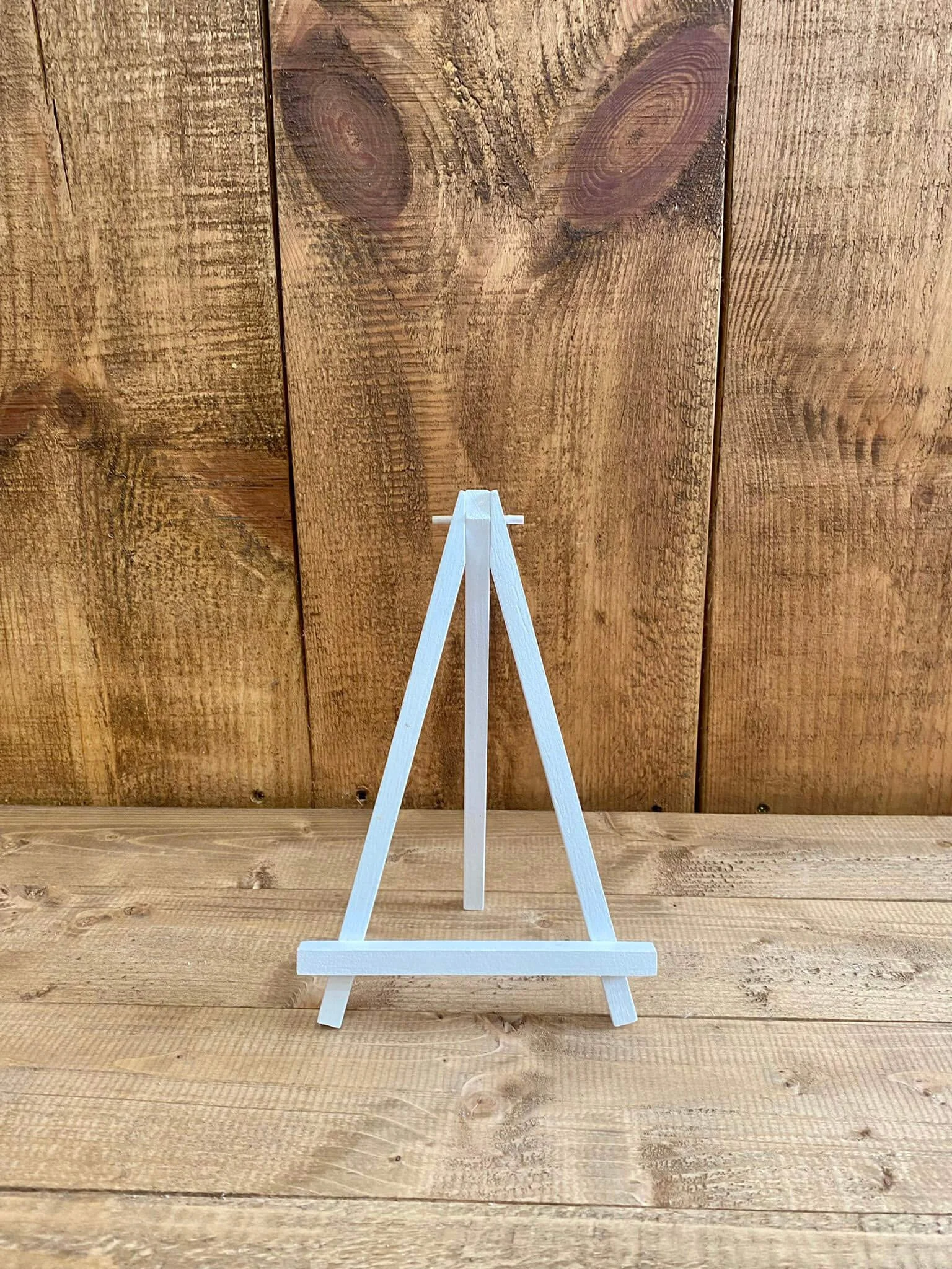 Mini Wood Easel - White standing on a wooden surface with a rustic wooden background, available to hire for your wedding or event.