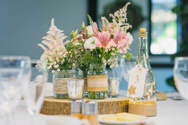 Rustic Jam Jar decorated with lace and twine, filled with pink and white flowers, available to hire for your wedding or event.