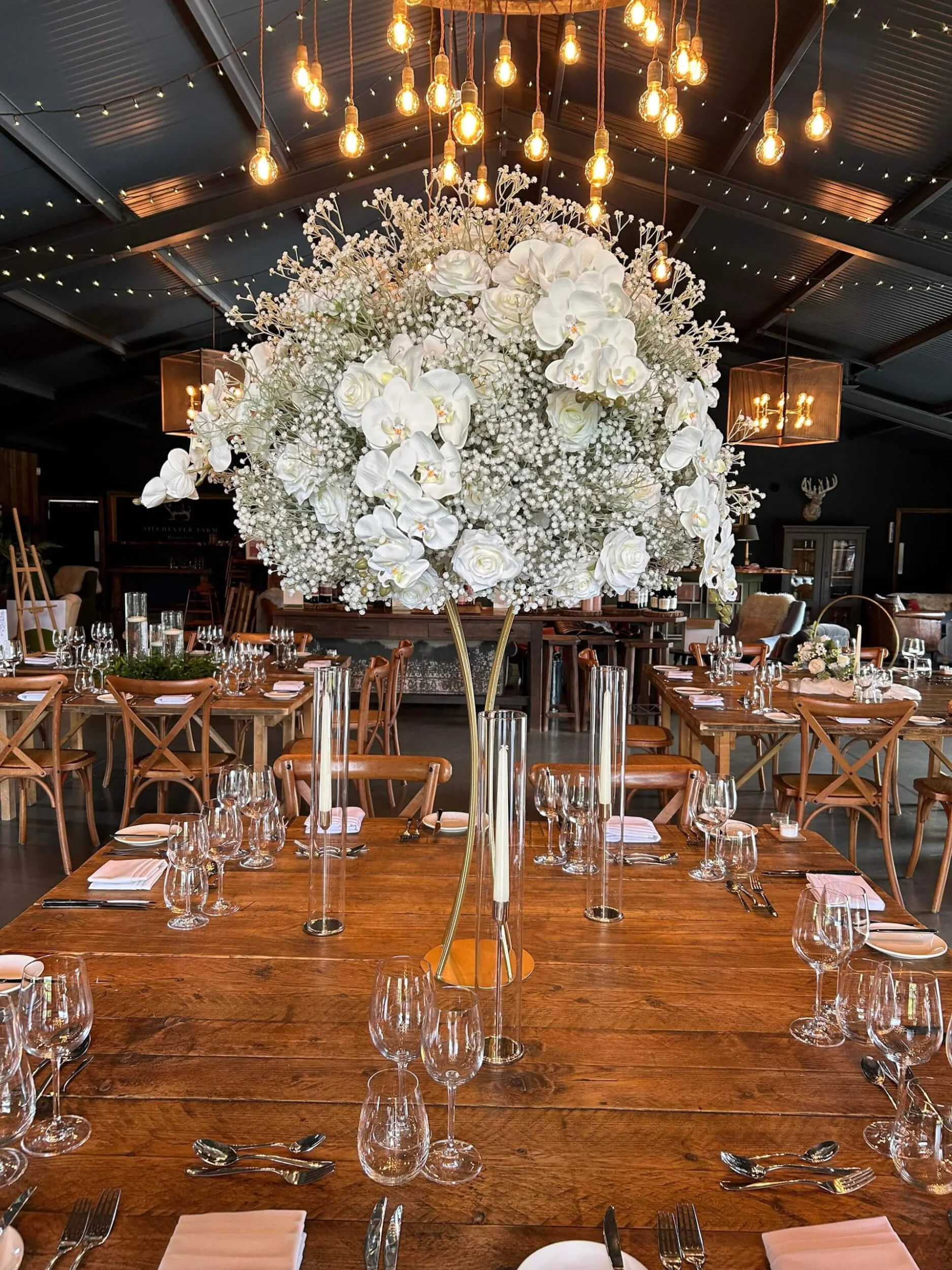 Lush white faux floral arrangement with orchids, roses, and baby's breath on a gold stand, available to hire for your wedding or event.