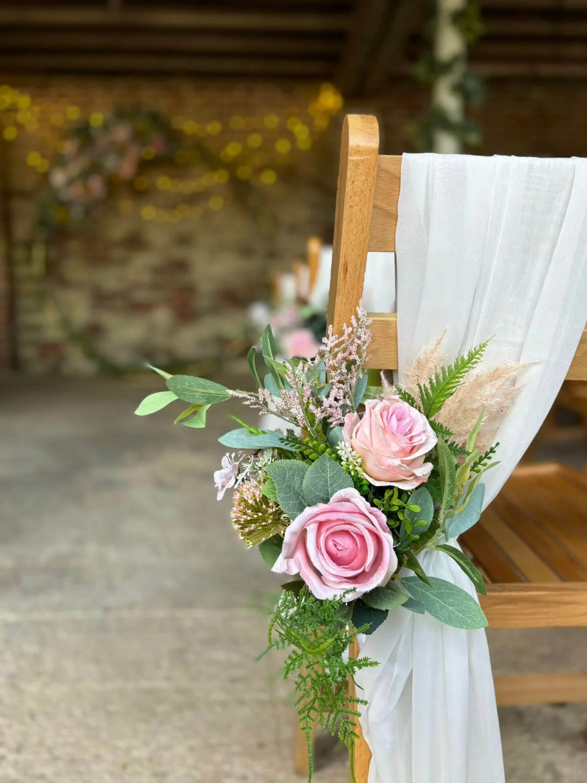 Faux Florals - Pew End faux floral arrangements bespoke colours with pink roses and greenery on a wooden chair draped in white fabric, available to hire for your wedding or event.