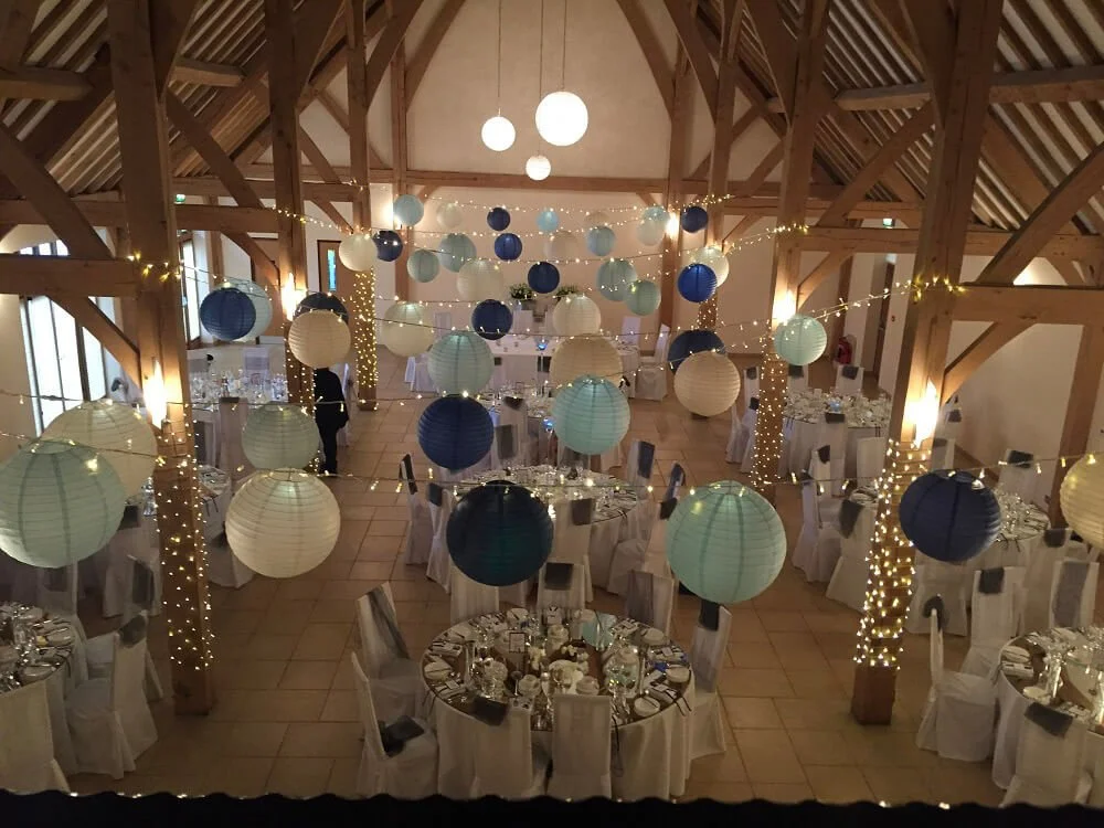 Hanging Lanterns Prices From in blue and white shades, strung across a wooden beam venue with fairy lights, available to hire for your wedding or event.