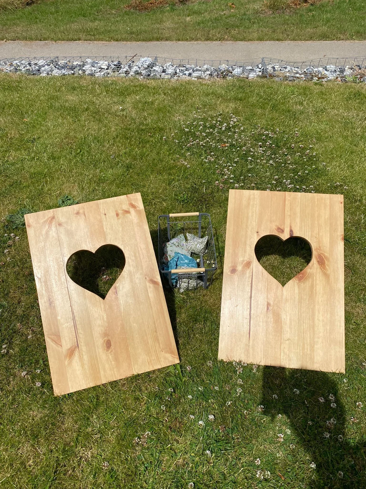 A rustic wedding garden game featuring two natural wooden cornhole boards with heart-shaped cut-outs and a basket of fabric bean bags. Ideal for outdoor weddings, garden parties, barn weddings and rel