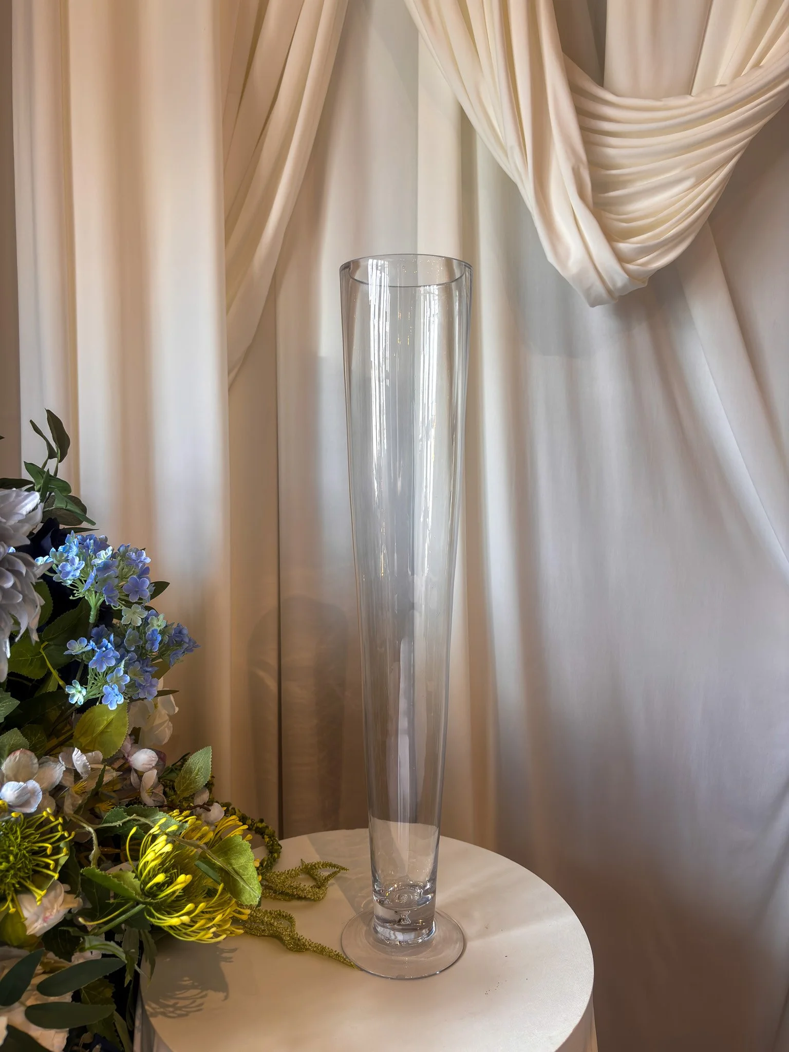 Hire our 60cm tall clear glass footed vase for weddings and events. Perfect for elegant floral centrepieces, with bespoke faux floral arrangements also available.