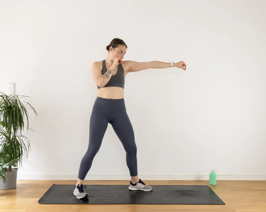A woman in workout clothes practicing boxing punches on a yoga mat in a bright room with a plant on the left and a water bottle on the right.