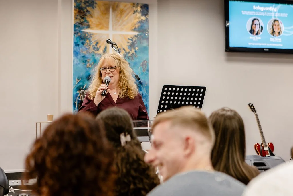 A woman with curly blonde hair and glasses speaking into a microphone during a presentation at an event, with a diverse audience in front of her.