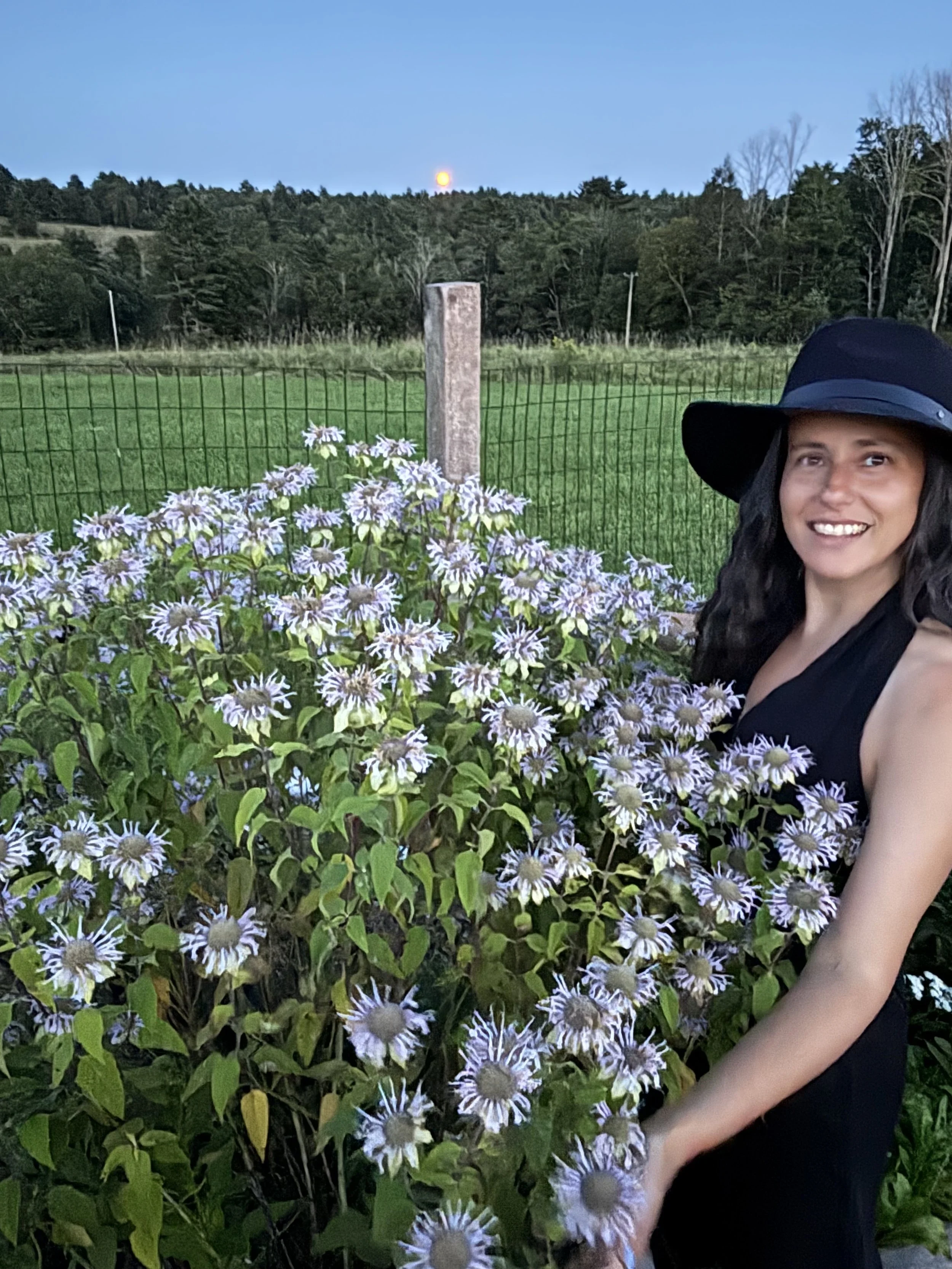 A woman with long dark hair, wearing a black wide-brimmed hat and black tank top, smiling in front of purple and white daisies in a garden, with a green grassy field, trees, and a partly cloudy sky in the background.