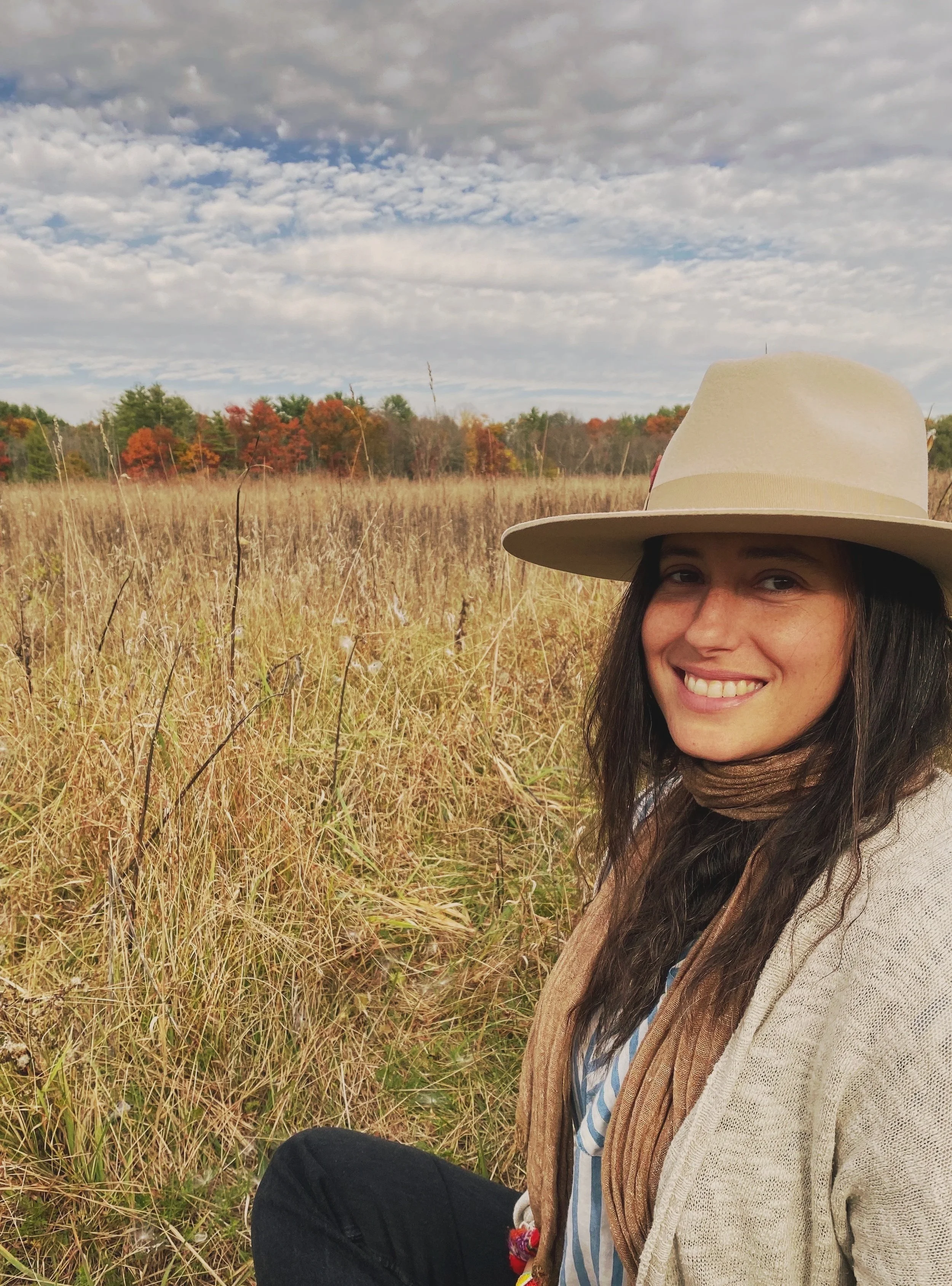 Smiling woman with long dark hair wearing a beige wide-brimmed hat, striped shirt, brown scarf, and beige cardigan, sitting in a field of tall, dry grass with trees showing fall foliage in the distance under a partly cloudy sky.
