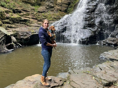Woman in hiking boots standing in front of a waterfall holding a small brown dog.
