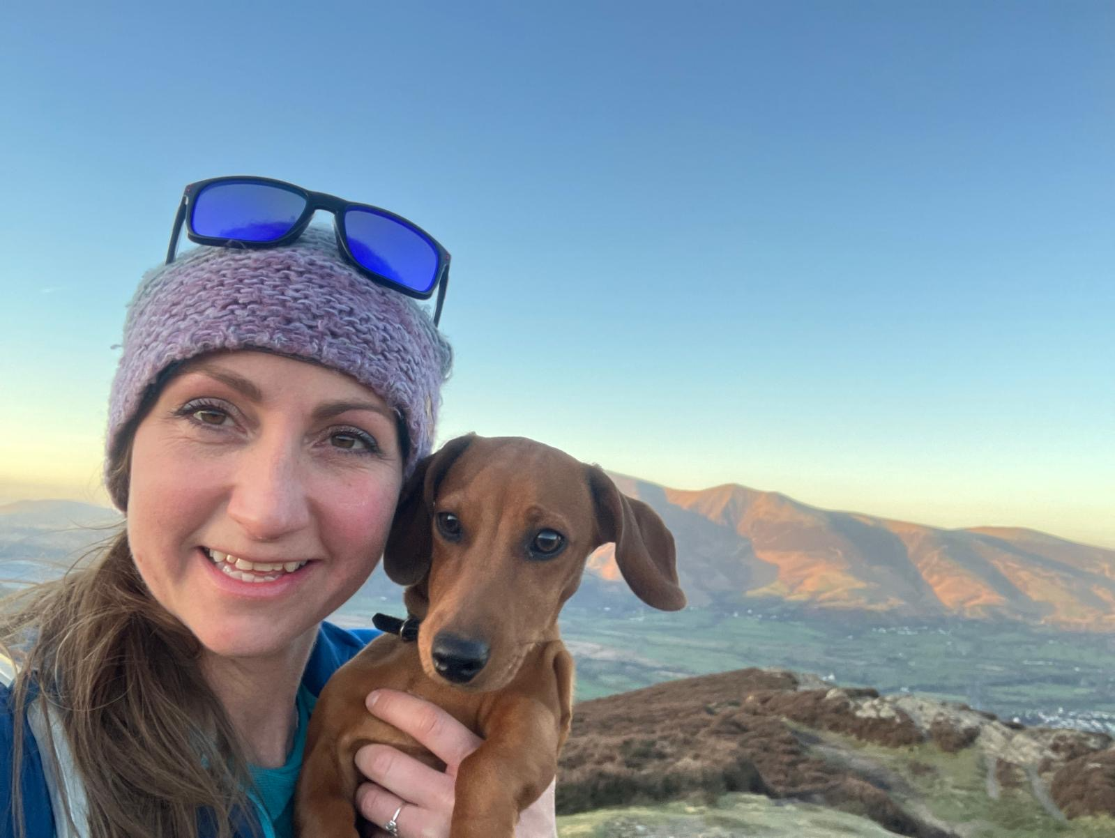 Woman with a wool hat and sunglasses on her head holding up a small brown dog with a view of hills behind them.