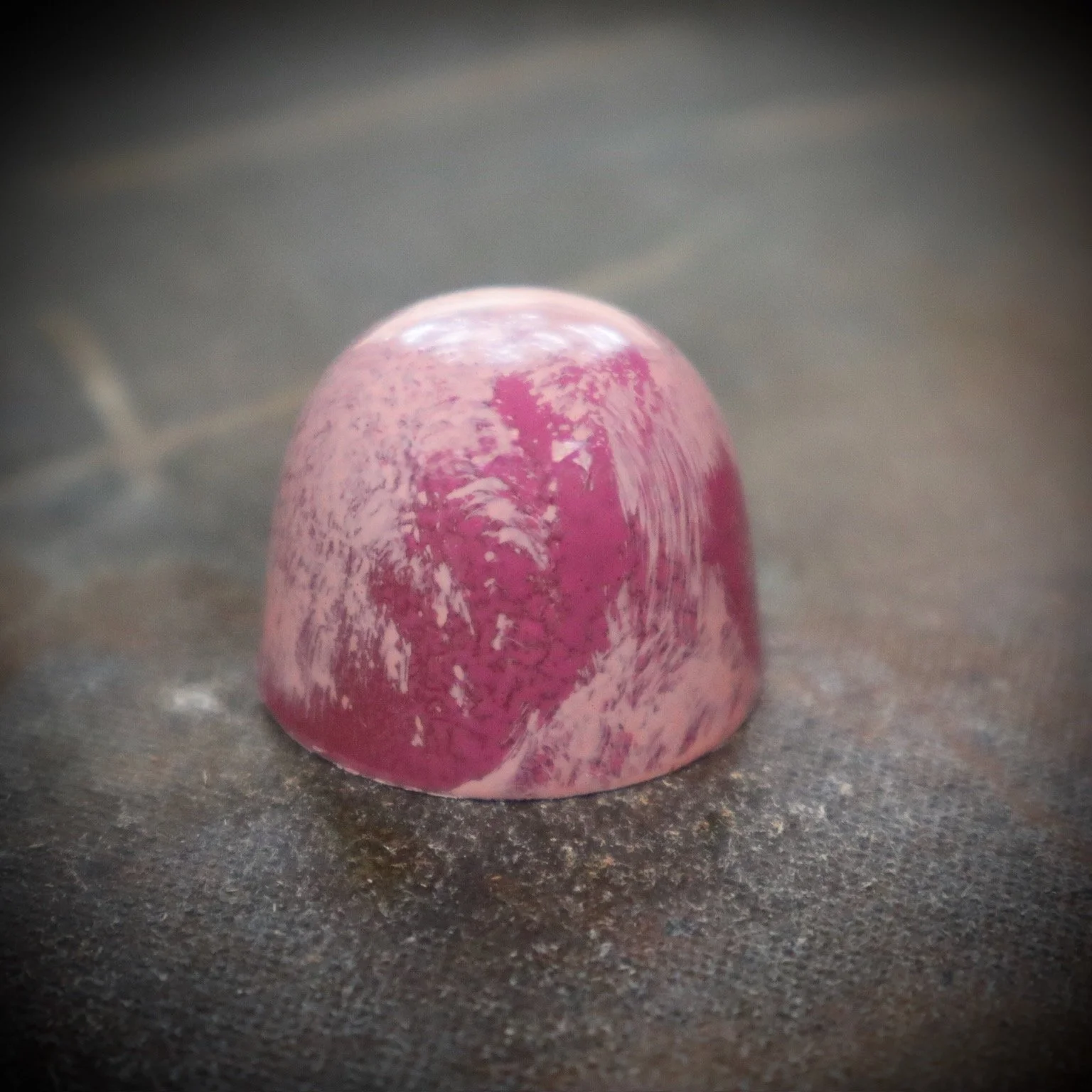 A pink and purple marbled, semi-glossy, cup-shaped piece of chocolate or confectionery on a dark textured surface.