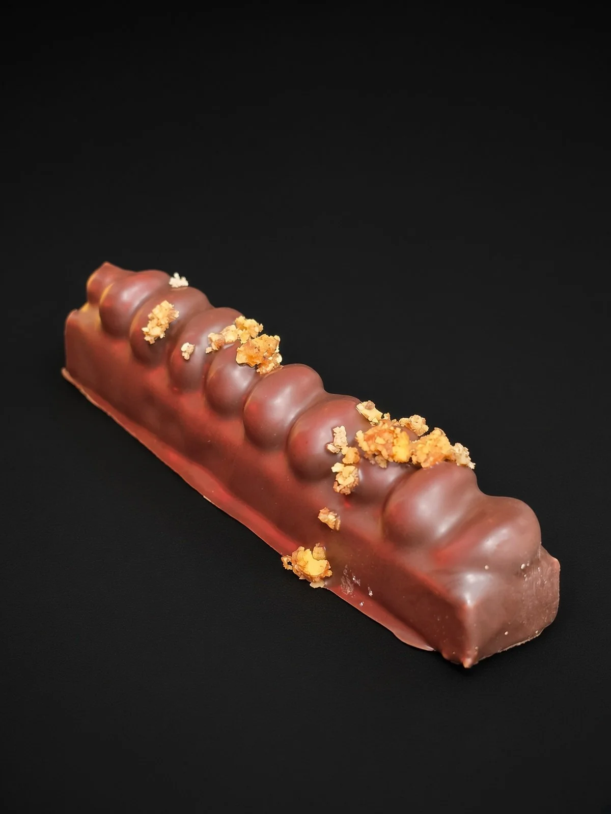 A long chocolate candy bar with a rippled, scalloped top scattered with crushed candied pecans, sharply in focus and well lit, resting on a solid matte black background.