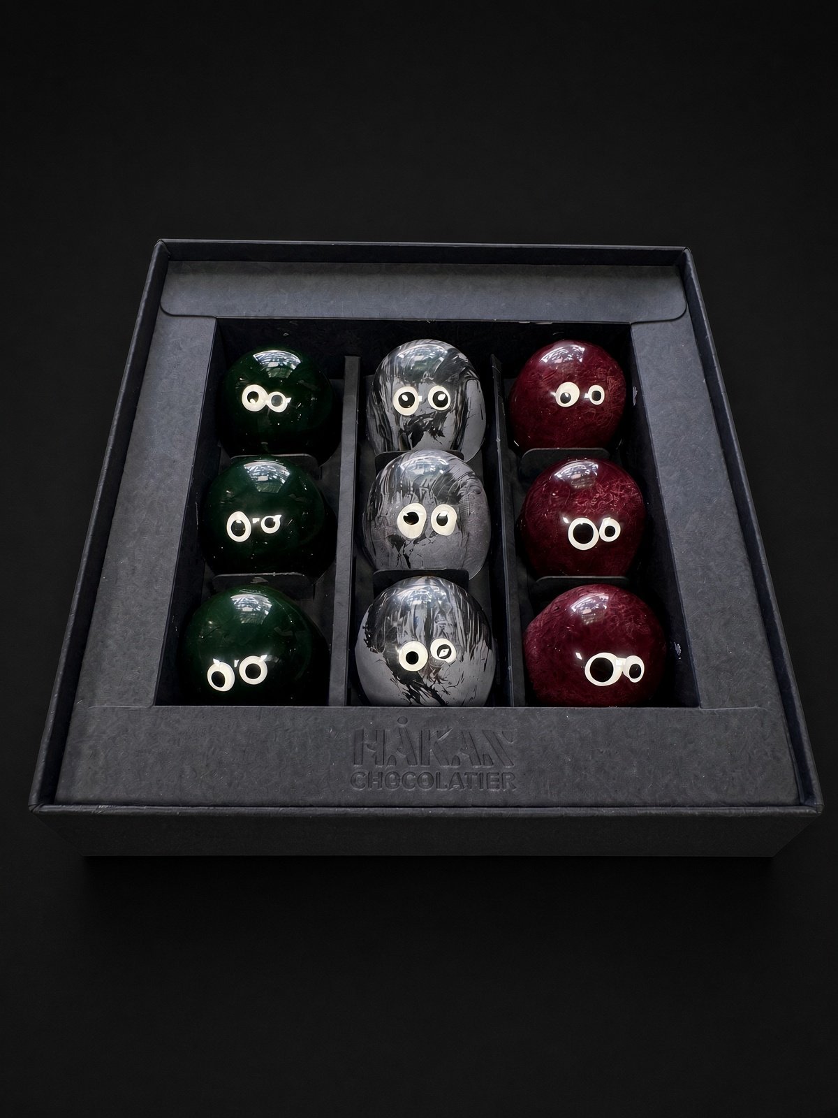 The 9-piece Little Monsters box: an open black HÅKAN box holding rows of green Frankie, gray Wolfie and deep red Dracula bonbons, each with candy eyes, on a dark background.