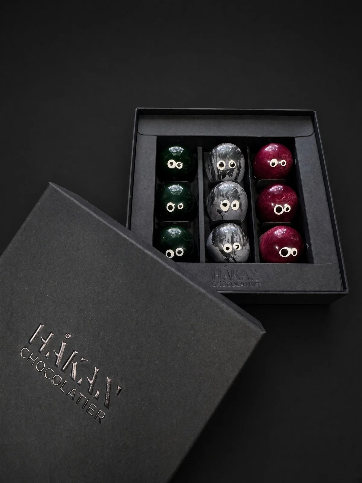 The 9-piece Little Monsters box, open beside its black HÅKAN Chocolatier lid, holding green, gray and deep red bonbons with candy eyes, on a dark background.
