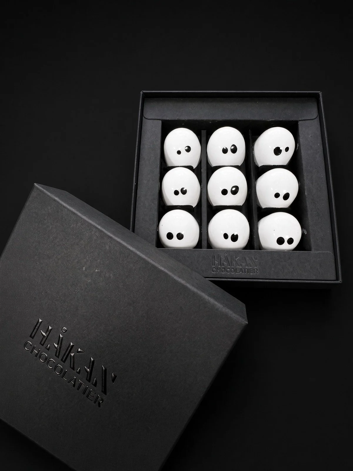 The Boo Box, open beside its black HÅKAN Chocolatier lid, holding nine white Ghost bonbons with black eyes, on a dark background.