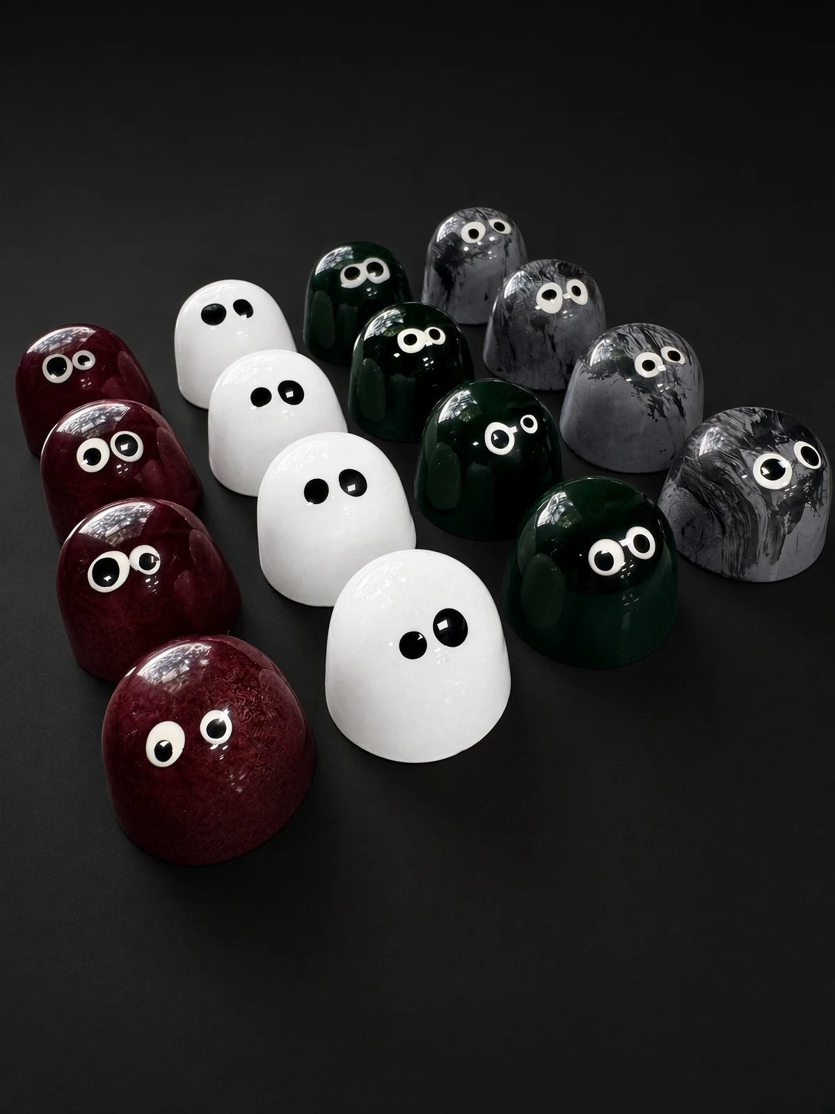 Four rows of Halloween bonbons with candy eyes: deep red Dracula, white Ghost, green Frankie and gray Wolfie, on a dark background.