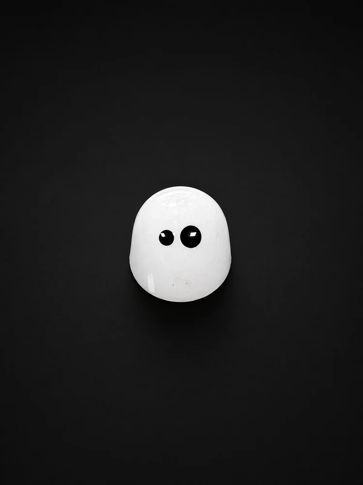 A glossy white dome-shaped chocolate bonbon with two black eyes, like a little ghost, sharply in focus and well lit, resting on a solid matte black background.