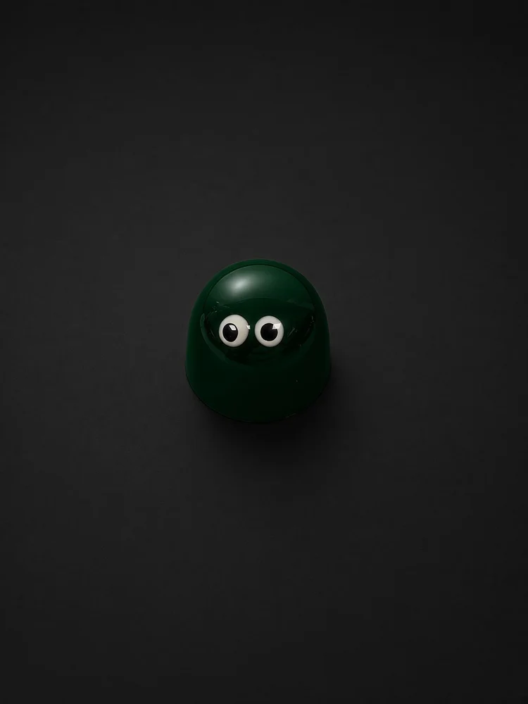 A glossy dark green dome-shaped chocolate bonbon with two white candy eyes, sharply in focus and well lit, resting on a solid matte black background.