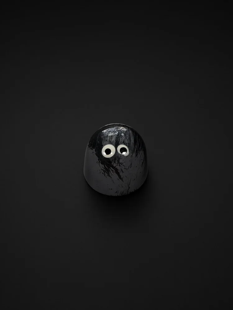 A glossy charcoal-gray dome-shaped chocolate bonbon with dark brushstroke streaks and two white candy eyes, sharply in focus and well lit, resting on a solid matte black background.