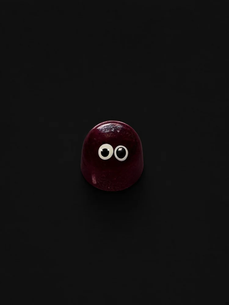 A glossy deep purple dome-shaped chocolate bonbon with two white candy eyes, sharply in focus and well lit, resting on a solid matte black background.