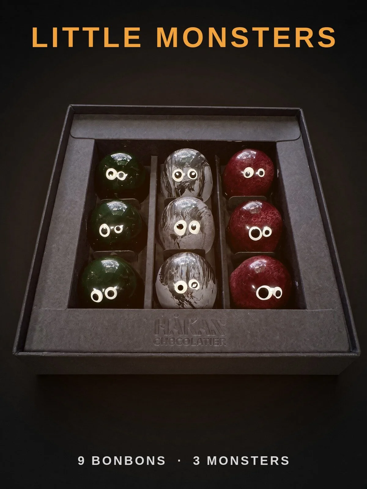 Little Monsters: an open black HÅKAN box of nine Halloween bonbons with candy eyes, green Frankie, gray Wolfie and deep red Dracula, on a dark background.