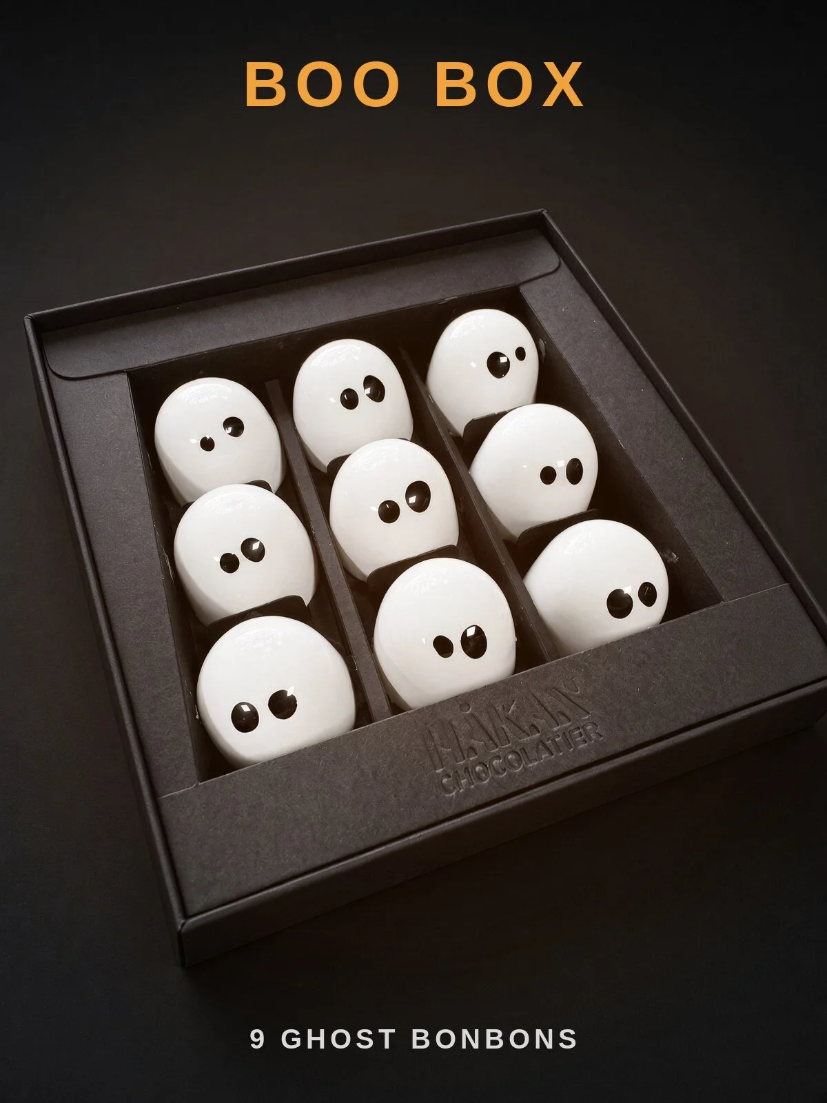 Boo Box: an open black HÅKAN box holding nine white Ghost bonbons with black eyes, on a dark background.