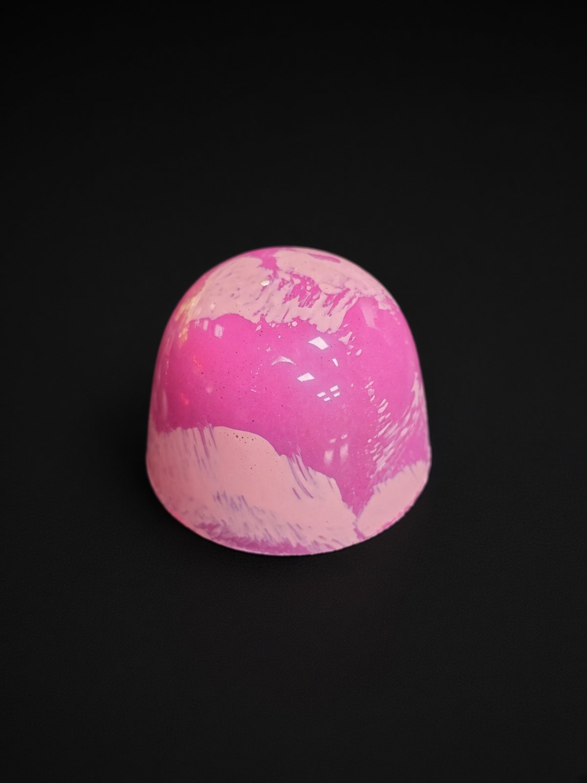 A glossy bright pink chocolate bonbon with soft pale-pink brushstroke swirls, sharply in focus and well lit, resting on a solid matte black background.