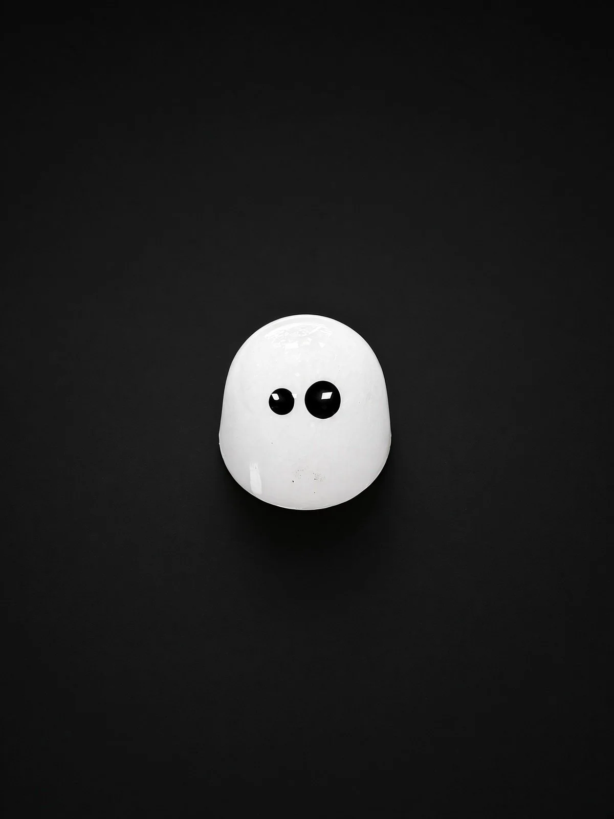 A glossy white dome-shaped chocolate bonbon with two black eyes, like a little ghost, sharply in focus and well lit, resting on a solid matte black background.