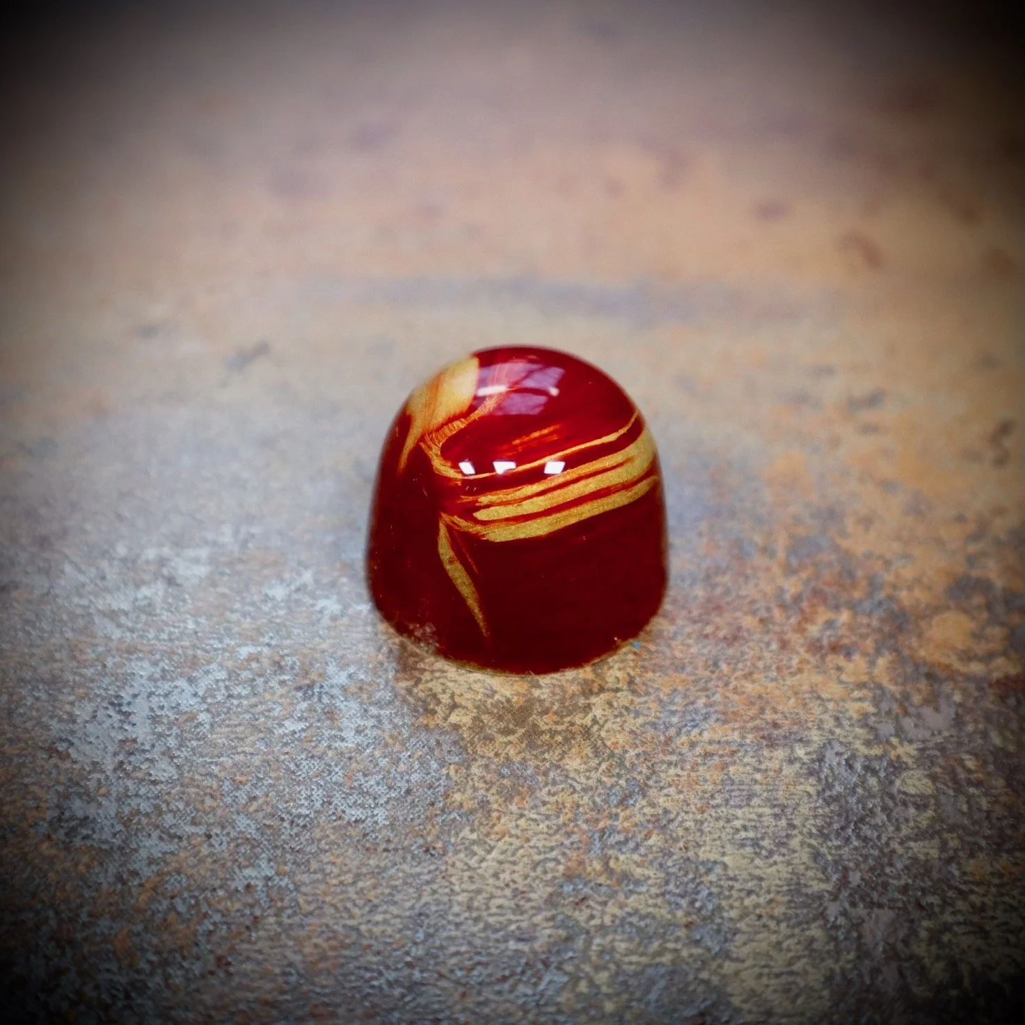 A polished, chocolate bonbon with red and yellow swirled patterns, sitting on a textured surface.