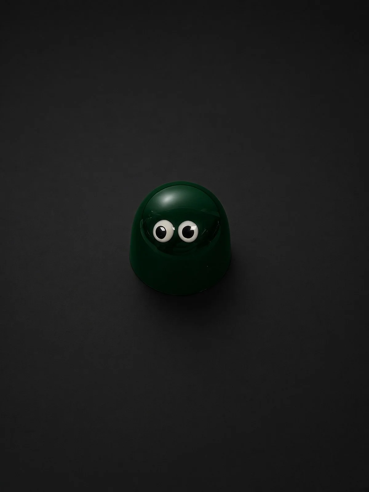 A glossy dark green dome-shaped chocolate bonbon with two white candy eyes, sharply in focus and well lit, resting on a solid matte black background.
