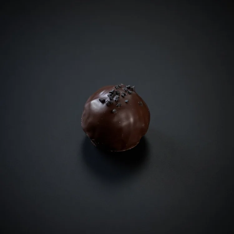 A Black Yuzu Kinetic Bomb: a glossy dark chocolate sphere topped with a sprinkle of black salt, resting on a solid matte black background.